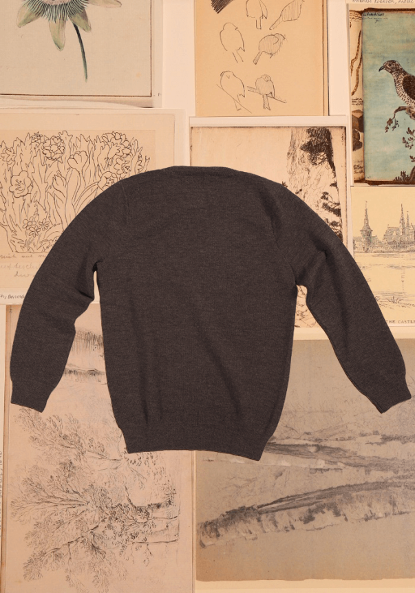 A charcoal gray, long-sleeve, crewneck sweater with ribbed cuffs and hem, displayed flat against a backdrop of vintage botanical and architectural sketches.