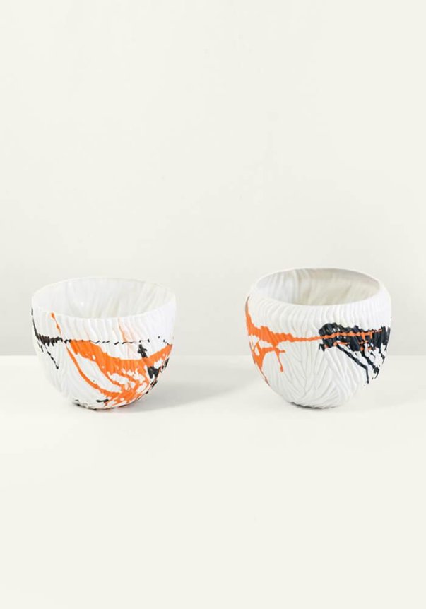Two textured white ceramic bowls with abstract orange and black splatter designs on a plain light background.