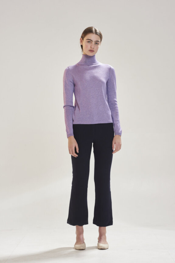 A person is wearing a light purple turtleneck and black pants, standing against a plain background.