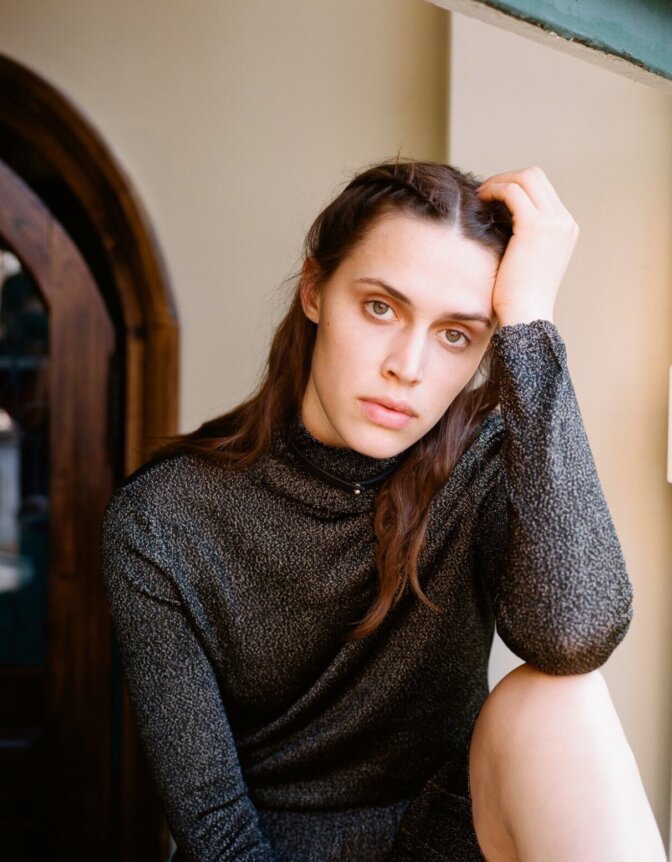 A person with braided hair and a thoughtful expression, wearing a gray long-sleeve top, sits by a window.