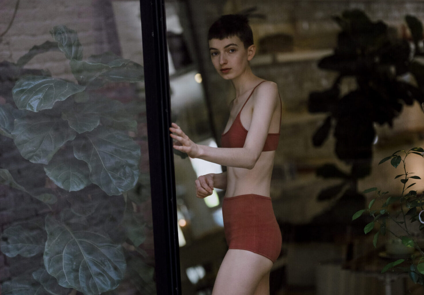 A person wearing a matching red bralette and high-waisted bottoms is standing by a window with plants visible inside and outside.