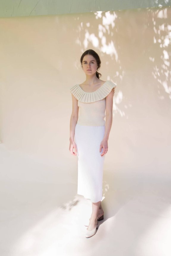 A woman is wearing a cream-colored sleeveless top with a large pleated collar paired with a fitted white midi skirt and low block heel shoes in a softly lit setting.