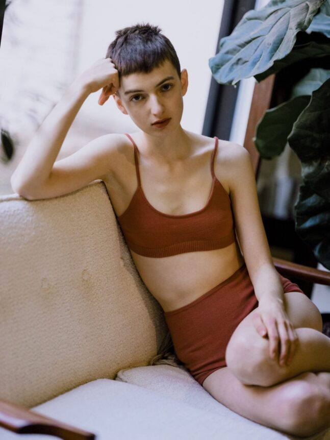 A person with short hair sits on a couch wearing a red bralette and shorts.
