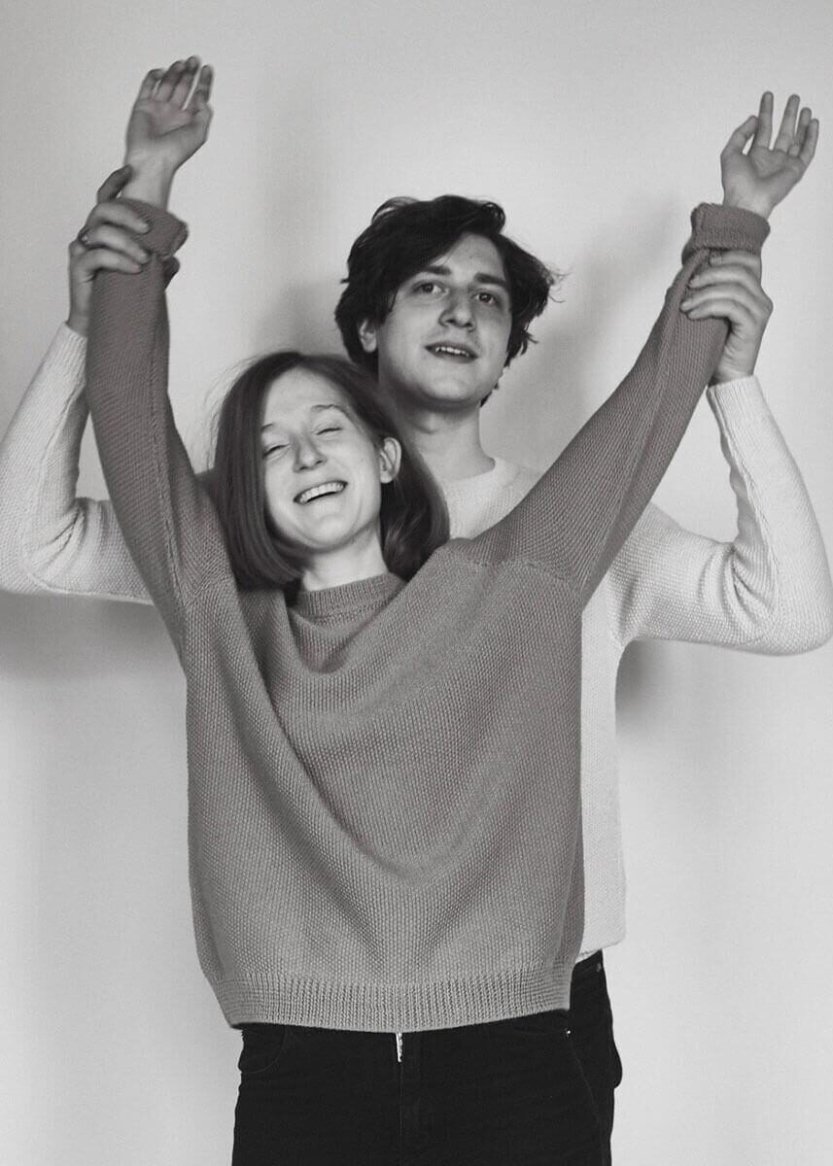 Two individuals wearing cozy, long-sleeve knit sweaters in soft, textured fabric, one in a warm shade of pink, the other in white, embracing joyfully.