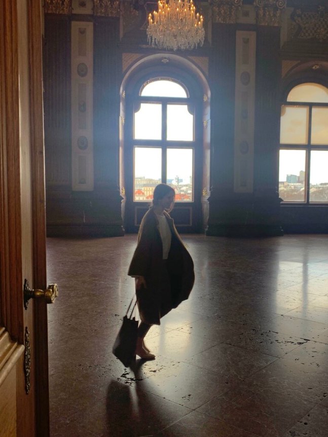 A person in a loose-fitting, brown wool coat and beige clothing carries a black tote bag in an opulent, chandelier-lit room with arched windows.