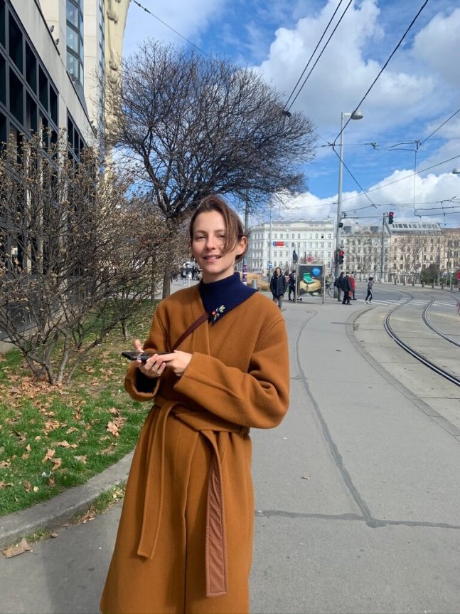 A woman is wearing a caramel brown belted overcoat with a relaxed fit, complemented by a black turtleneck, detailed with a floral brooch, against an urban backdrop.