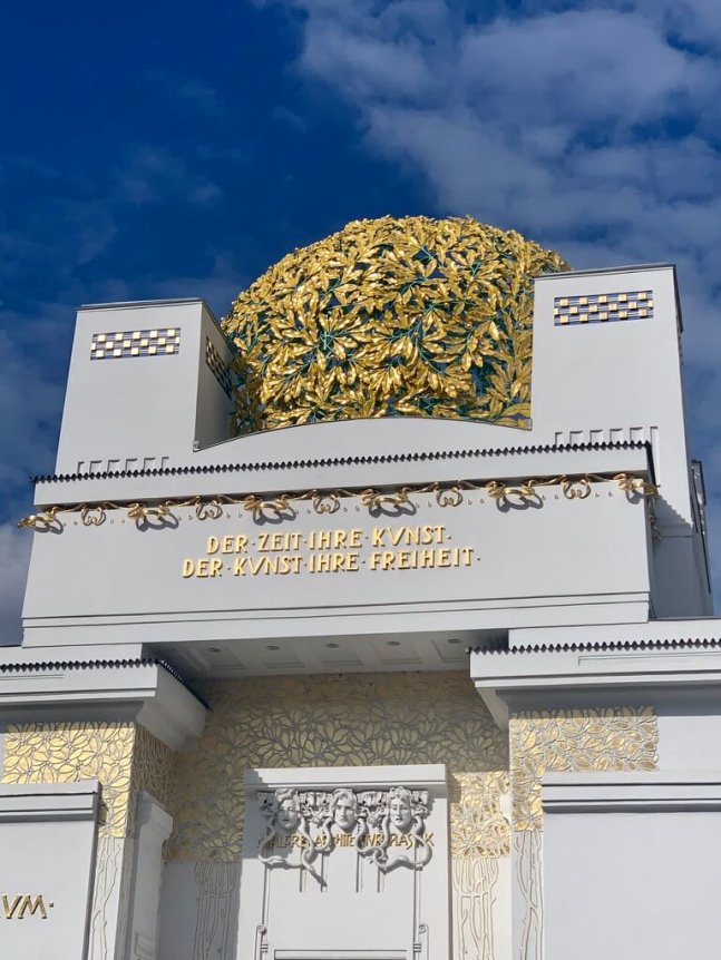 The image showcases a white building with a golden ornamental dome and decorative elements against a clear blue sky.