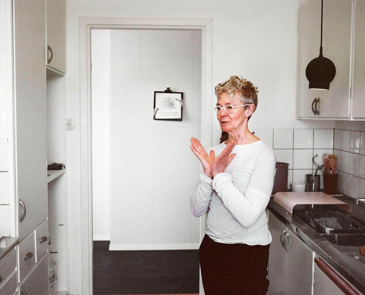 A woman in a white, long-sleeved, semi-sheer top with a loose fit stands in a minimalist kitchen.