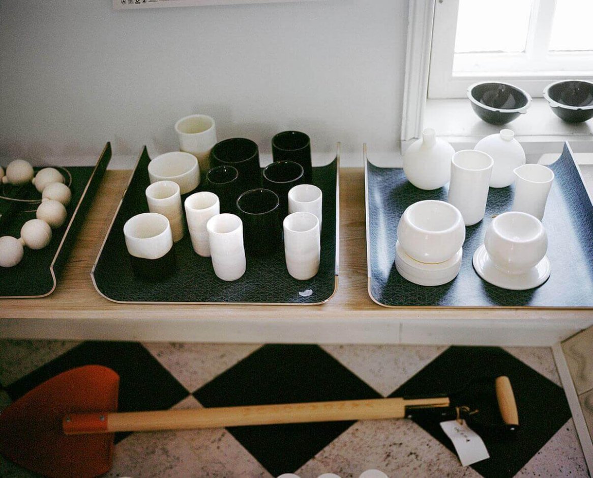 The image features a collection of black and white ceramic cups and vases displayed on trays, accompanied by a small shovel on a checkered floor.