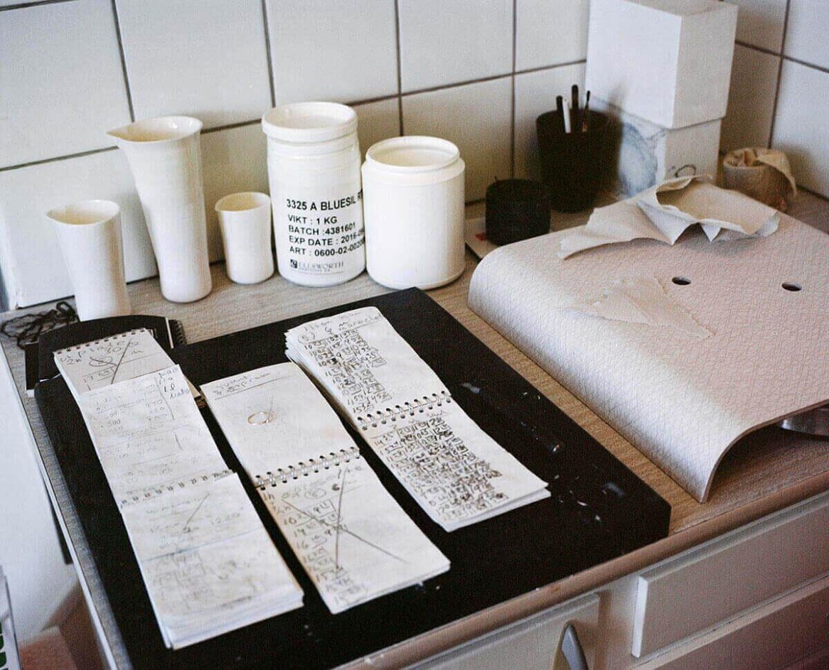 A workspace with white measuring cups, a container labeled "Blues" powder, notebooks filled with handwritten notes, and textured paper on a countertop.