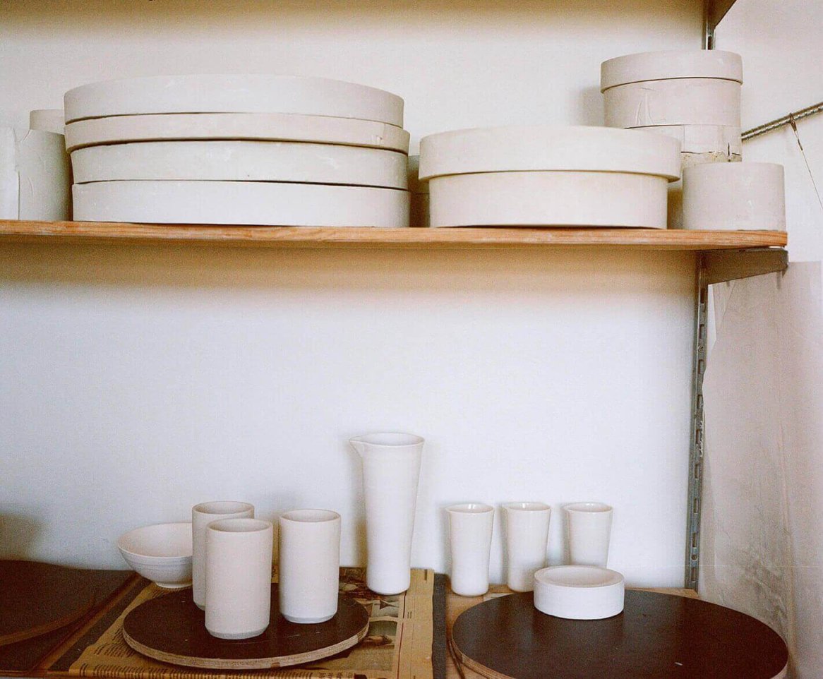 The image shows a collection of white ceramic molds and vessels on wooden shelves against a plain wall.