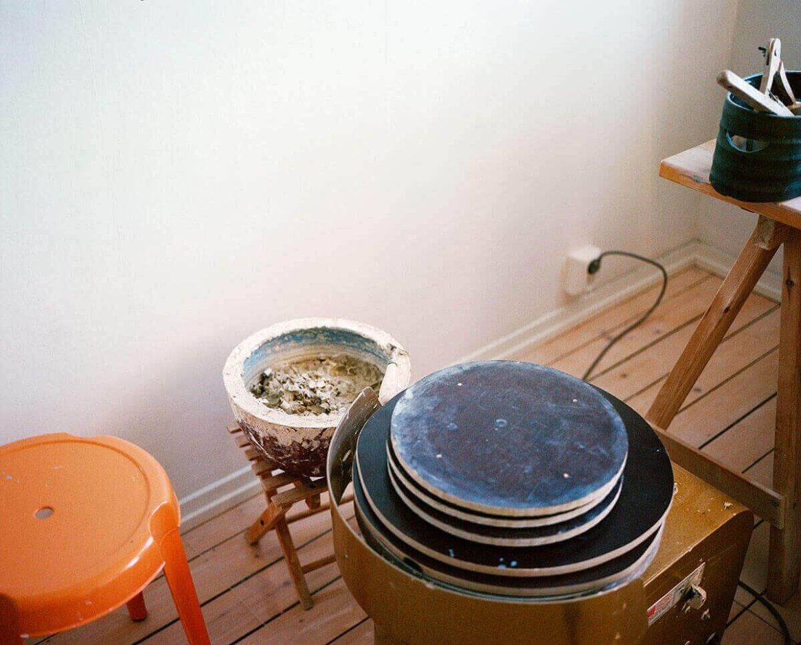 A pottery studio with a potter's wheel, an orange stool, and a workspace with ceramic tools on a wooden floor.