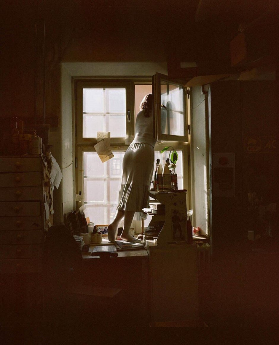 A woman in a fitted white knit top and skirt, standing on a desk by a window in a dimly lit, cluttered room.