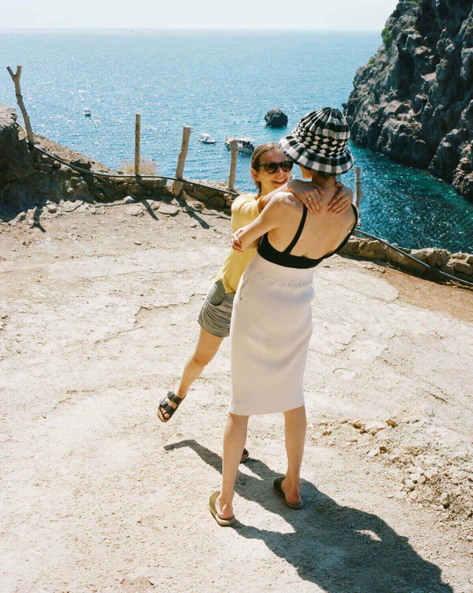 Two people embrace near a scenic ocean view, one in a fitted black tank top, textured white skirt, and patterned bucket hat, the other in a casual yellow t-shirt, gray shorts, and black sandals.