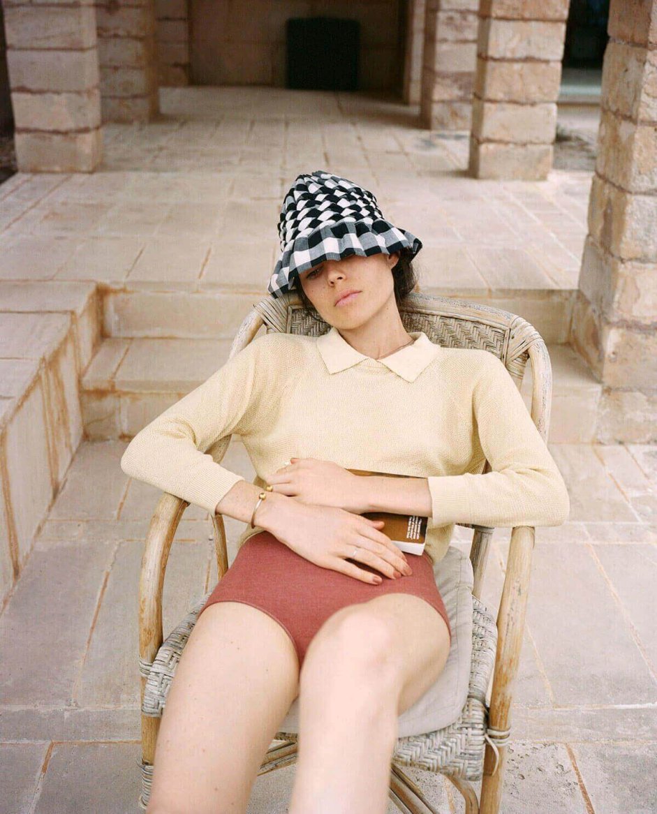 A model lounges in a relaxed-fit, cream-colored knit sweater with a collared neckline, paired with high-waisted, rust-colored shorts, accessorized with a checkered black and white bucket hat.
