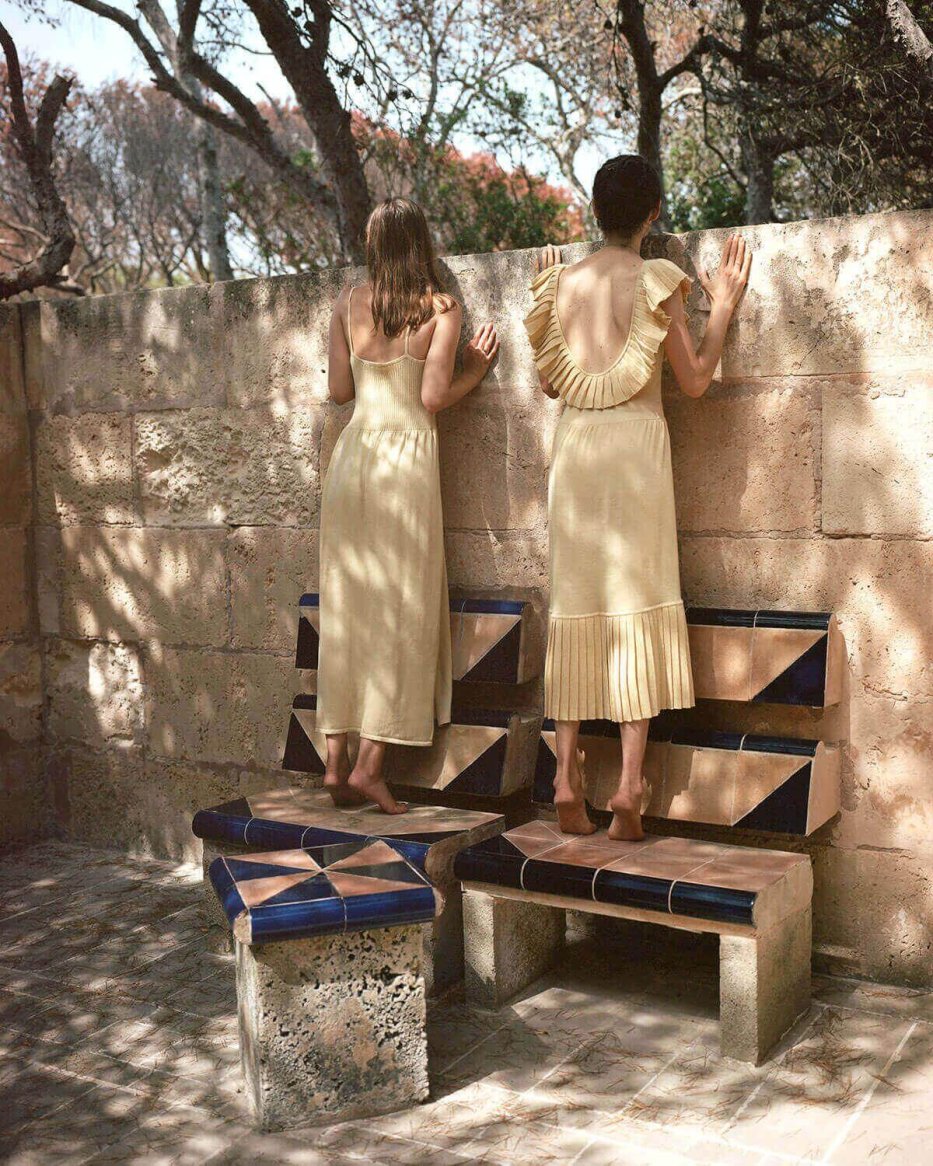Two women wearing light yellow, sleeveless, ankle-length dresses with knit texture and pleated details stand on mosaic-tiled benches, peering over a stone wall.