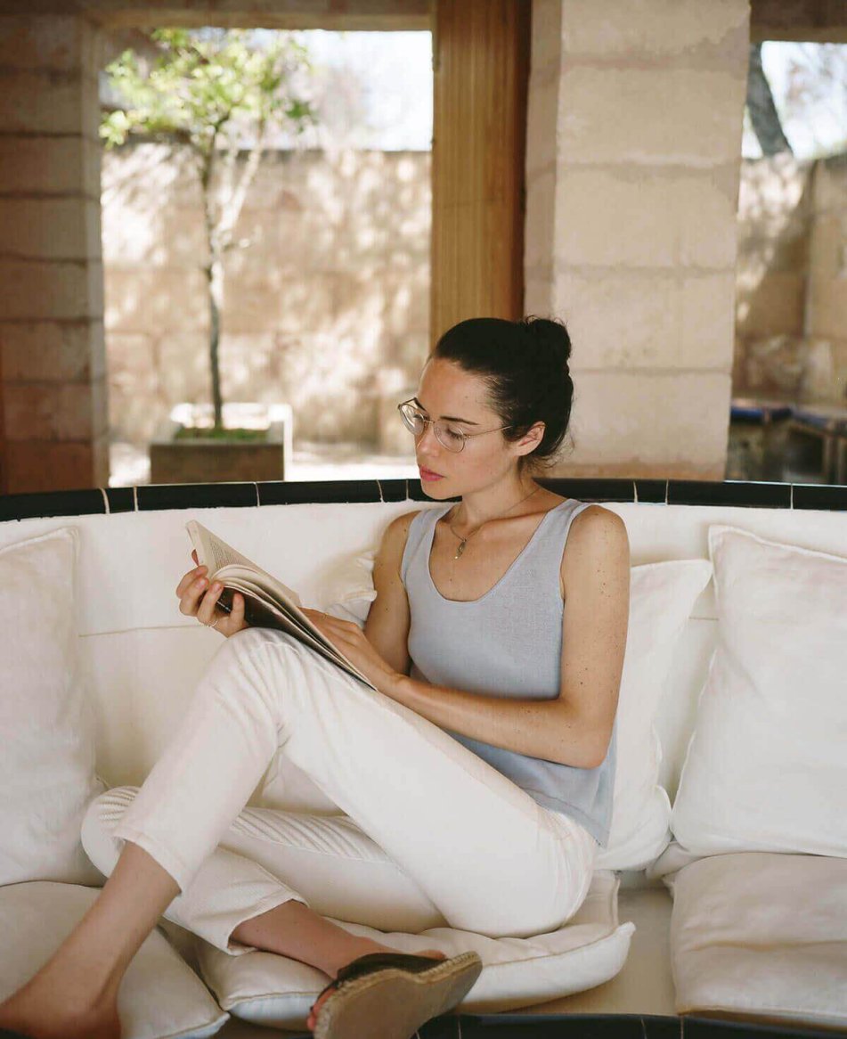 A woman is wearing a fitted, light blue sleeveless top and white slim-fit pants, complemented by glasses, reading on a white cushioned sofa.