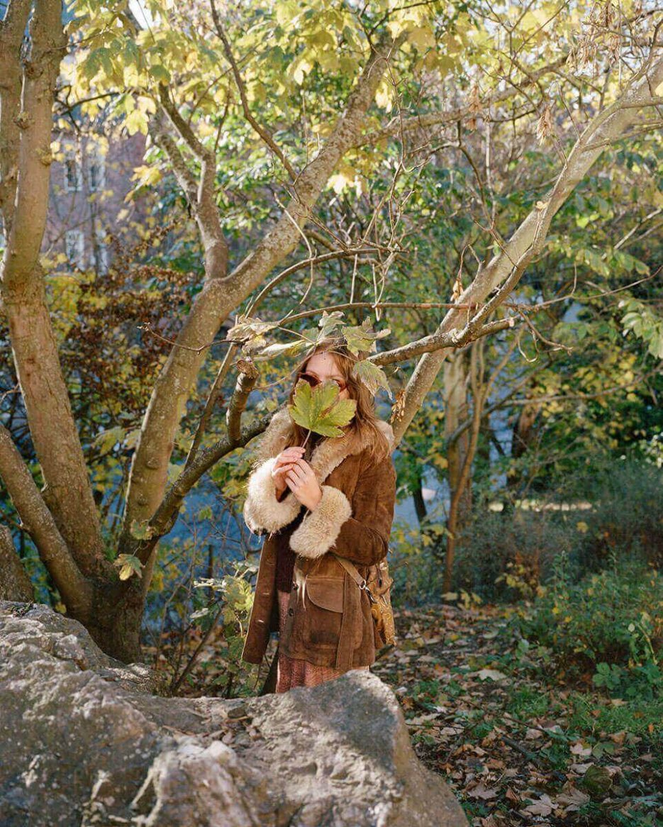 A model wearing a vintage brown suede coat with plush fur trim and large pockets stands amidst an autumnal forest backdrop, holding leaves.