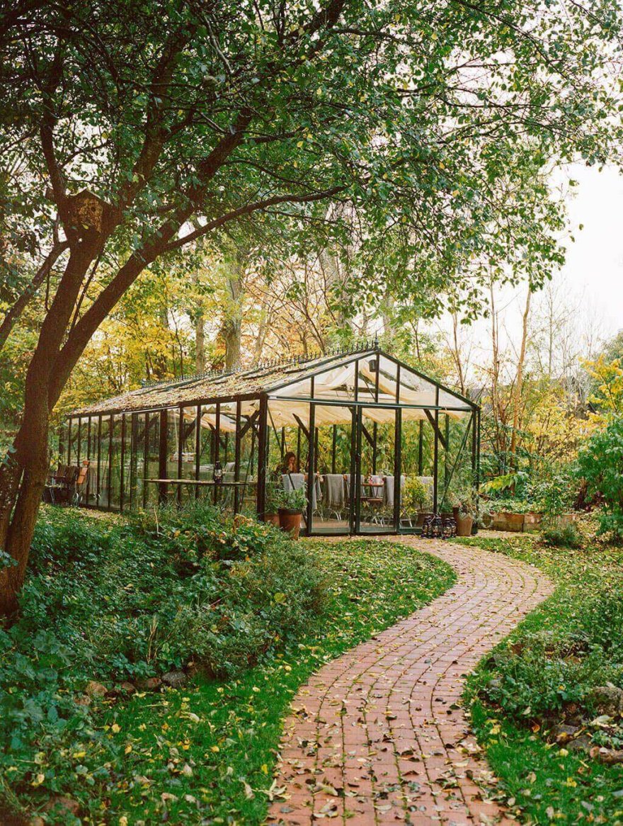 A quaint glass greenhouse surrounded by lush greenery and a winding brick pathway, with tables and chairs inside for relaxation or dining.