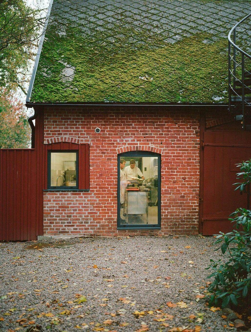 A rustic brick building with a moss-covered roof features a glimpse of chefs working inside, surrounded by a gravel courtyard and autumn foliage.
