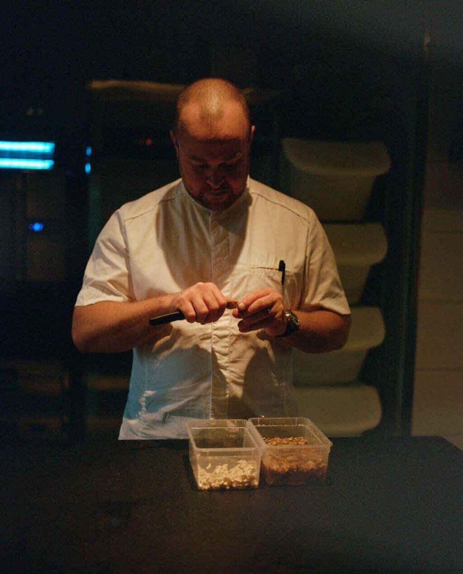 A man in a white, short-sleeve, button-up chef's shirt stands at a counter with two clear containers of ingredients.