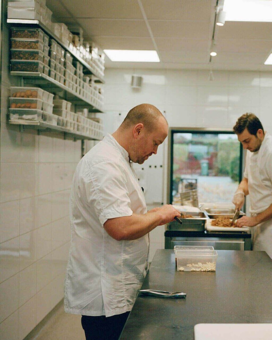 Two chefs in white short-sleeved chef jackets work in a professional kitchen with stainless steel surfaces and organized shelves.