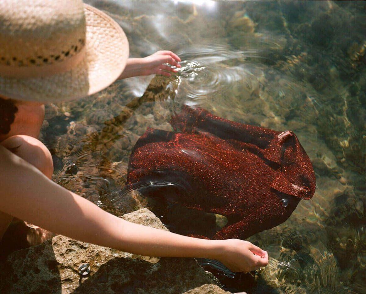 A shimmering, deep-red sequined top rests partially submerged in clear water, surrounded by rocks, with a person wearing a straw hat reaching towards it.