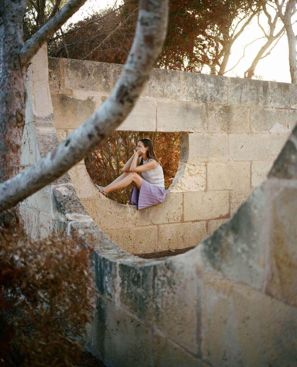 A woman in a lightweight, sleeveless white top and a loose-fitting purple skirt sits within a stone structure, surrounded by rustic trees.