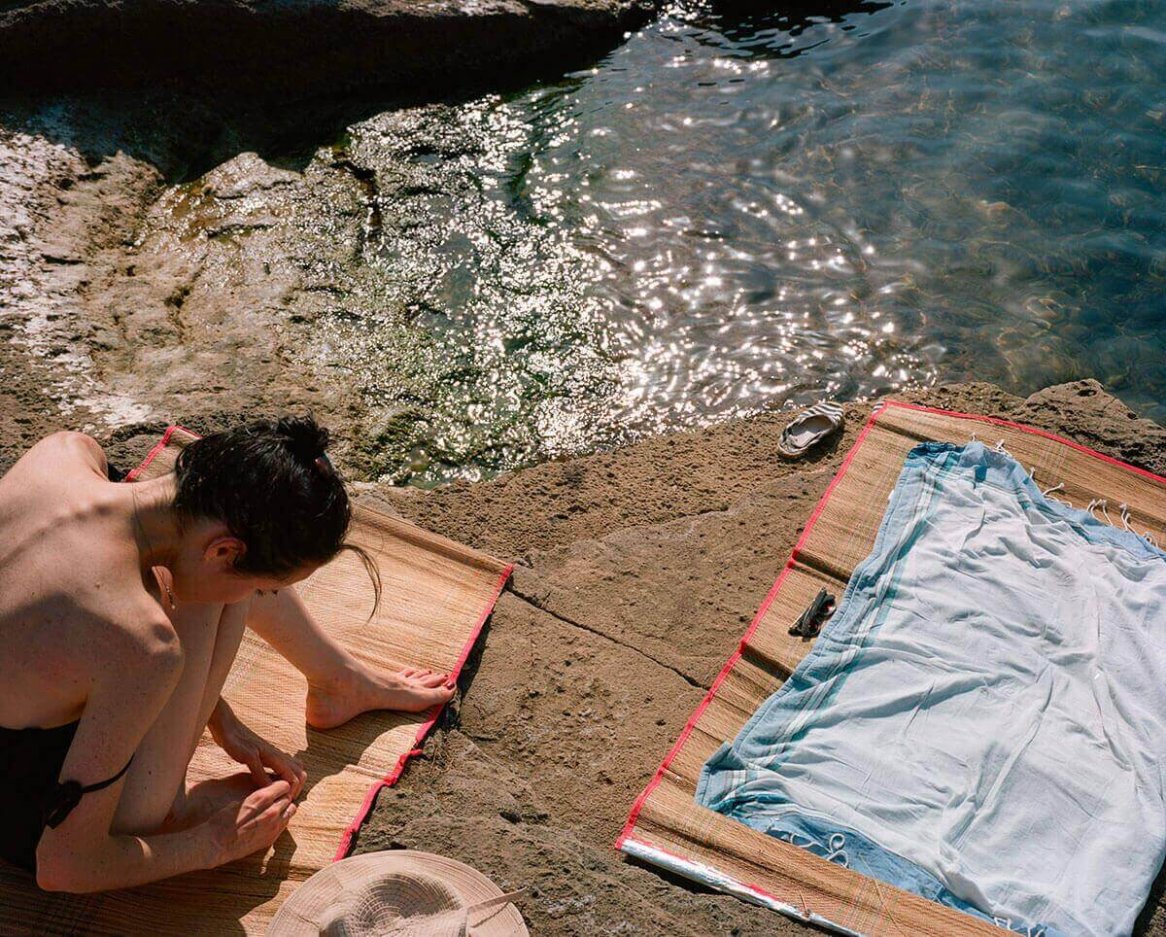 A person sits on a natural-colored woven mat near water, wearing a black swimsuit and accompanied by a beige woven hat on a rocky surface with sparkling water nearby.