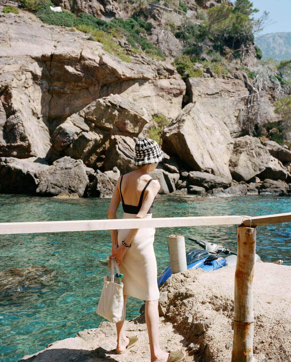 A person in a black top and cream knit skirt, accessorized with a checkered bucket hat and beige tote bag, stands by a rocky coastline with clear turquoise water.