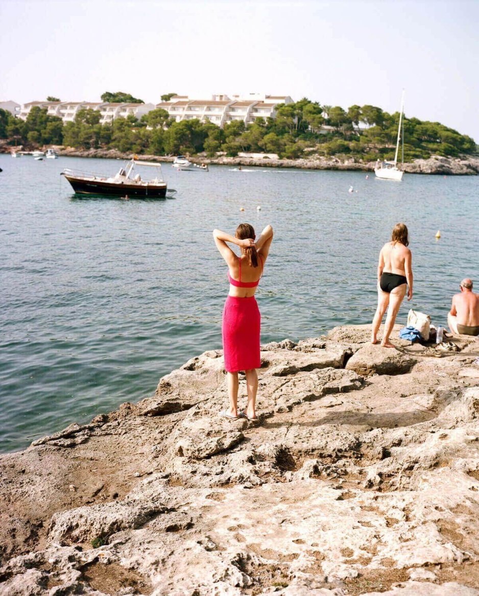 A woman in a fitted, bright red knit ensemble consisting of a crop top and high-waisted skirt stands on rocky terrain near the sea.