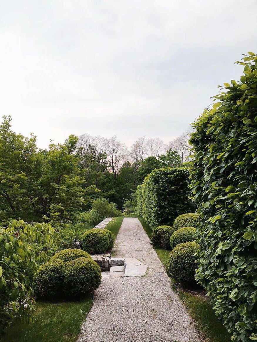 A neatly trimmed garden path lined with lush green hedges, round shrubs, and bordered by a gravel walkway under a cloudy sky.