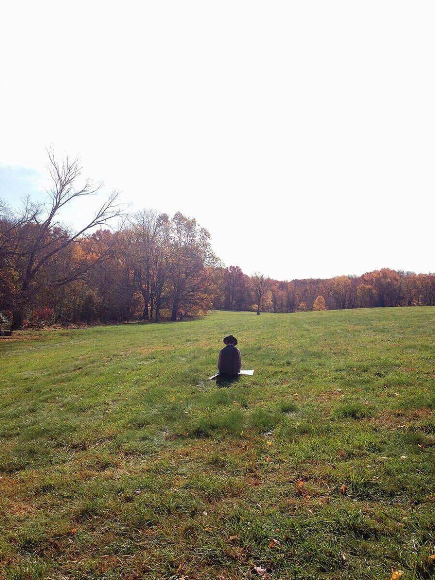 A person sits on a grassy field wearing a dark-colored, wide-brimmed hat and a loose-fitting coat, surrounded by autumn trees.