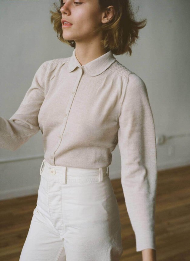 A woman wears a beige, long-sleeve, collared blouse with a textured finish paired with high-waisted, white pants featuring a tailored fit.