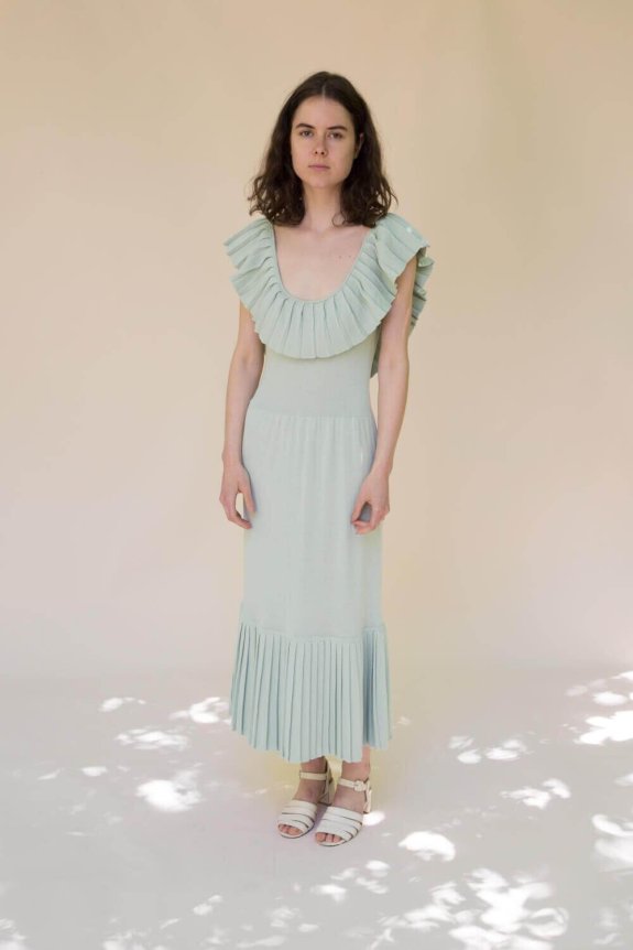 A model wears a light green, pleated, sleeveless dress with a ruffled neckline and fitted silhouette, paired with white sandals.
