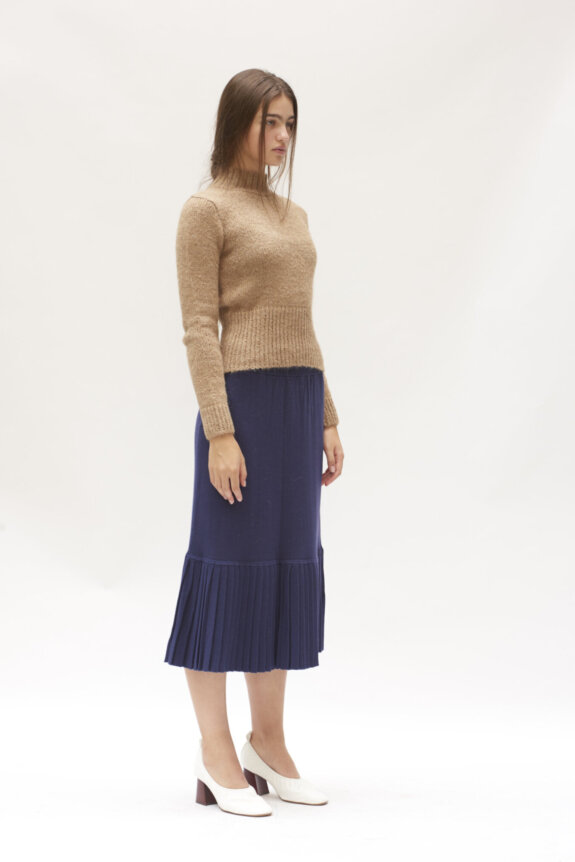 A woman in a beige sweater and blue pleated skirt stands against a plain background.