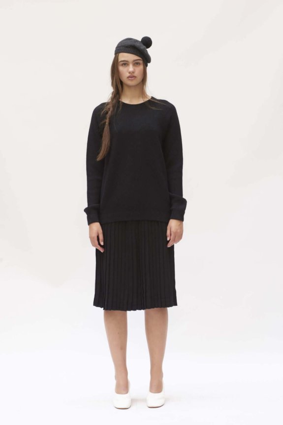 A model wears a black, relaxed-fit knit sweater with a pleated midi skirt, paired with a pom-pom beanie and white shoes.