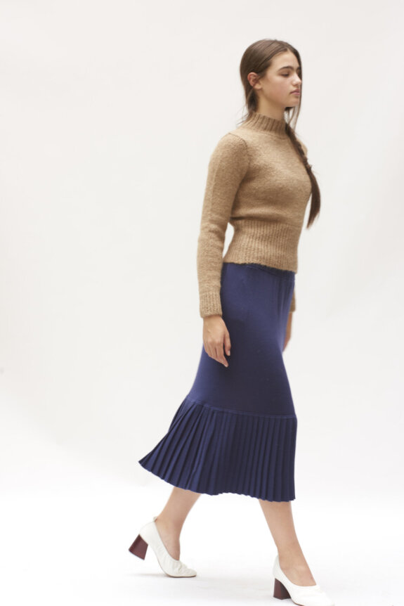 A woman in a tan sweater and blue pleated skirt walks in white heels.