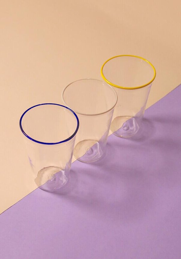 Three clear glasses with colored rims—blue, clear, and yellow—arranged on a split backdrop of beige and purple surfaces.