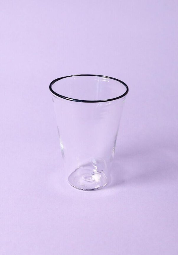 A clear glass tumbler with a black rim on a lavender background.