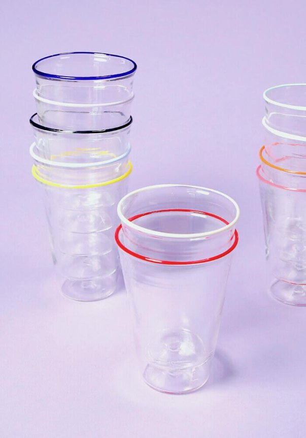 A set of transparent drinking glasses with colorful rims, stacked on a light purple surface.