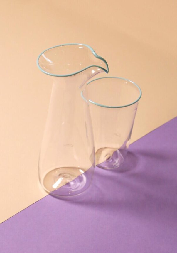 A clear glass pitcher and tumbler with a light blue rim, set against a beige and purple geometric background.