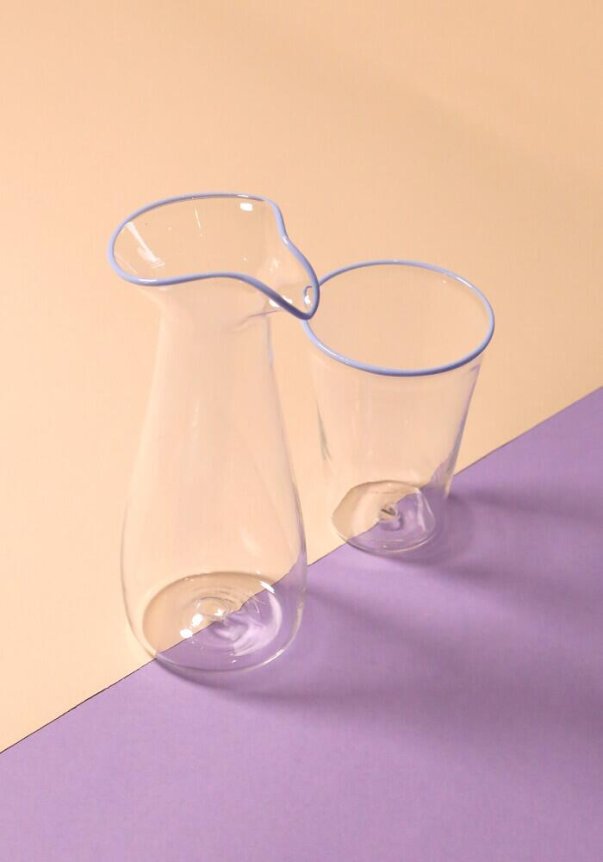 A transparent glass jug and cup with a light blue rim, placed on a dual-tone beige and lavender surface.