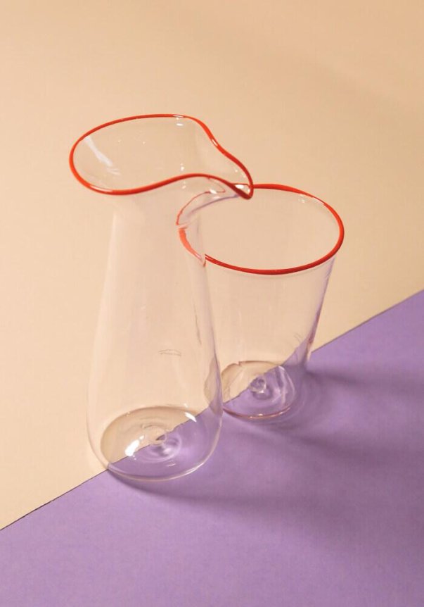 A clear glass pitcher and matching cup with red rims, set against a beige and lavender abstract background.