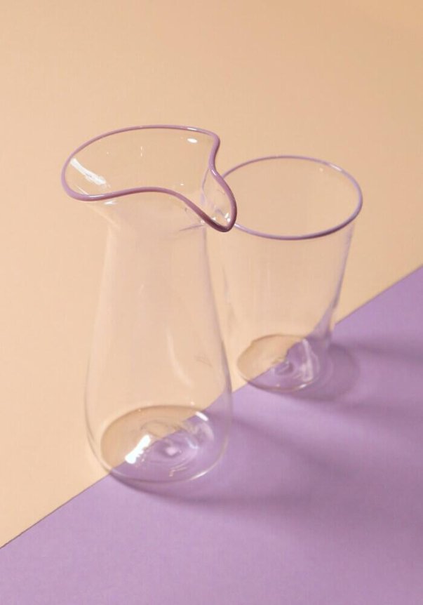 A clear glass carafe and cup with purple-rimmed edges sit on a peach and lavender-colored surface.