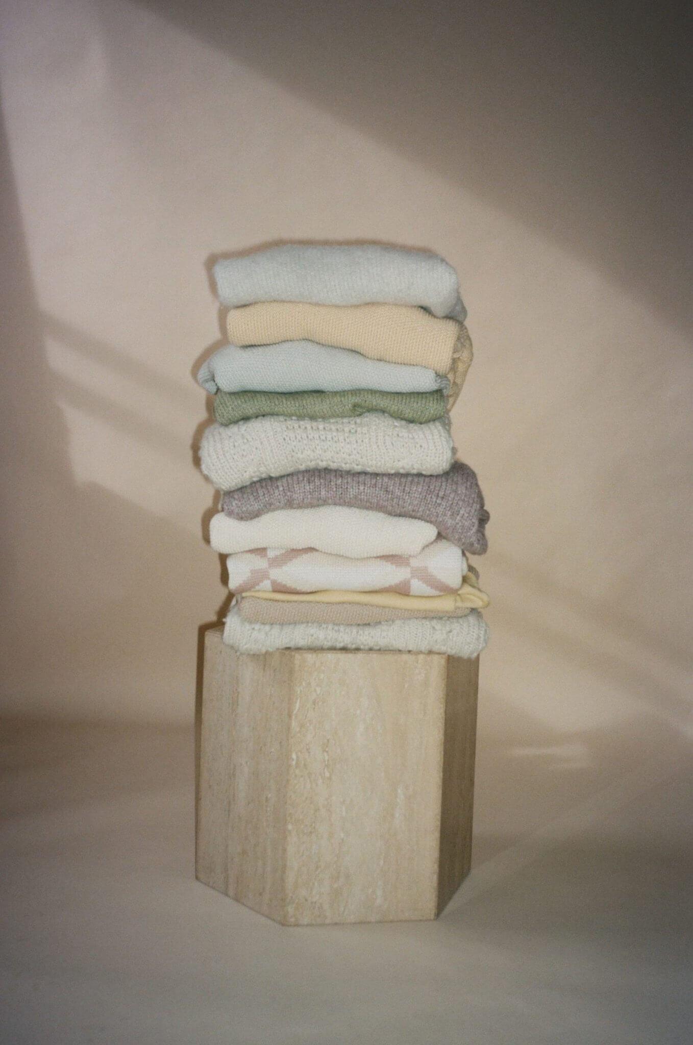A stack of pastel and neutral-colored knit sweaters, folded neatly on a hexagonal wooden pedestal against a soft, neutral background.