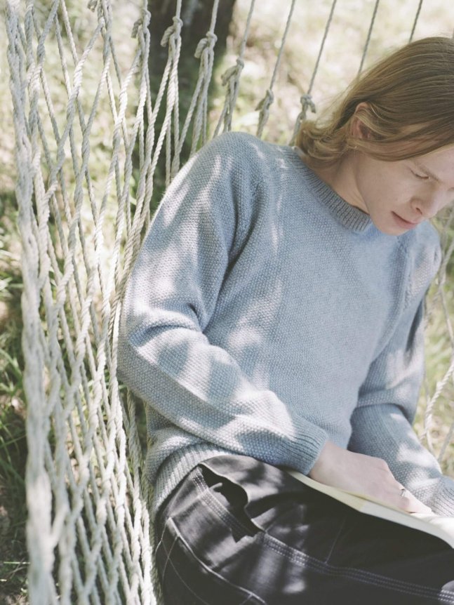 A person is wearing a light blue, loose-knit sweater with a relaxed fit, paired with dark pants, while sitting on a hammock outdoors.