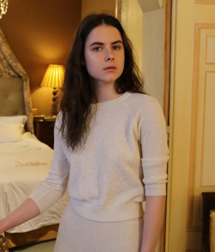A woman wearing an ivory, form-fitting, textured knit sweater and skirt ensemble in a luxurious bedroom setting.