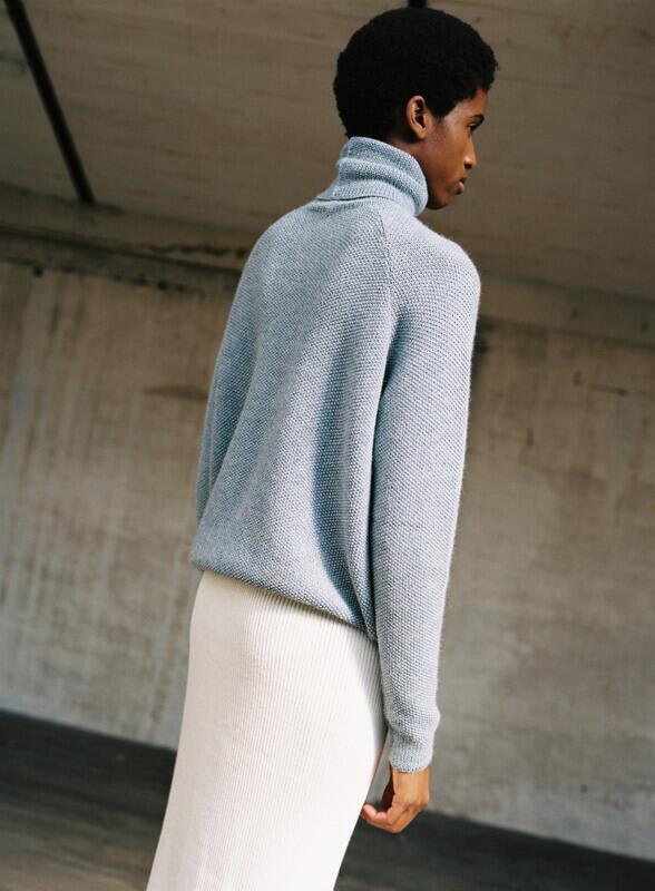 A model wearing a light blue, knit turtleneck sweater with a relaxed fit, paired with a white, ribbed skirt.