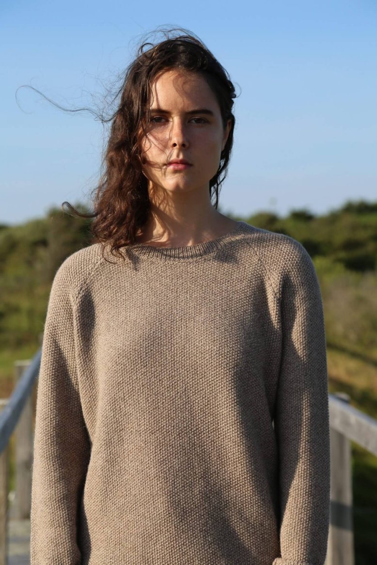 A woman wearing a loose-fitting, textured beige sweater made of a knit material, set against a natural outdoor backdrop.